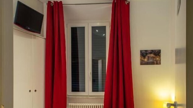 A bedroom with a bed, red curtains, a TV mounted on the wall, and a window with blinds. - Intrà residenza in trastevere (Rome)