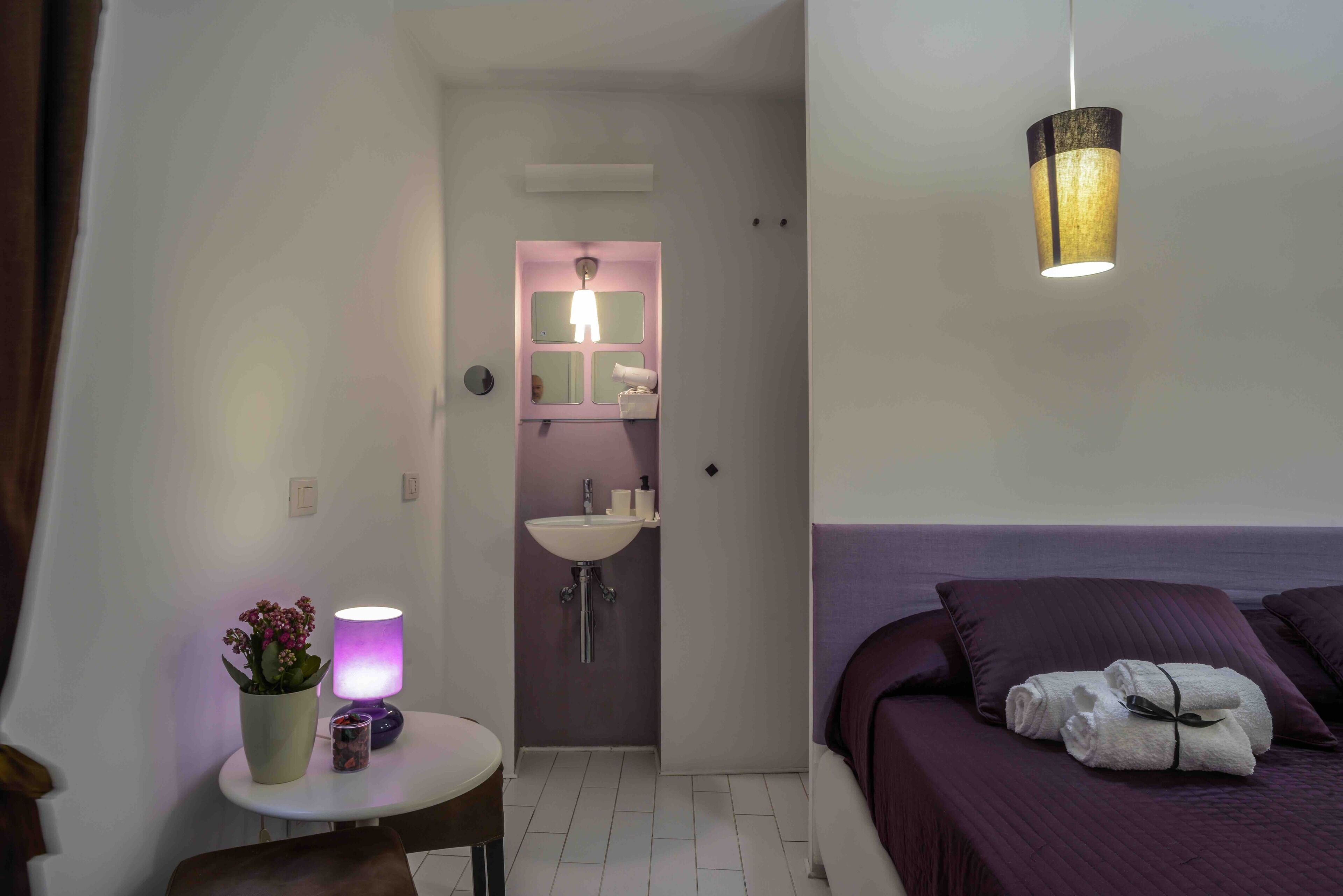 A hotel room with a bed, a bedside table with a lamp, a potted plant, and a bathroom visible through an open door.