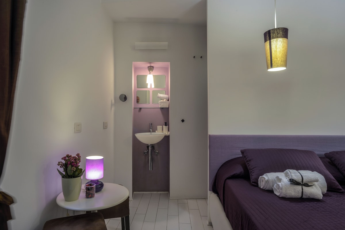 Une chambre à coucher avec un lit, une table de chevet avec une lampe, une plante en pot, et une salle de bain avec un lavabo et un miroir.