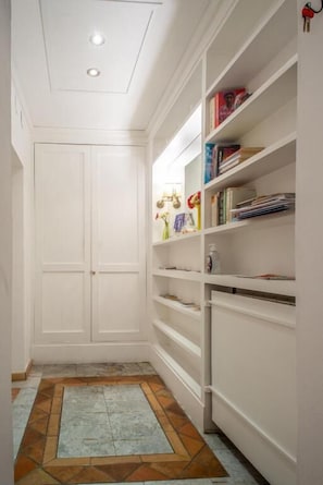 A narrow hallway with a tiled floor, white walls, and a built-in bookshelf filled with books and decorative items.