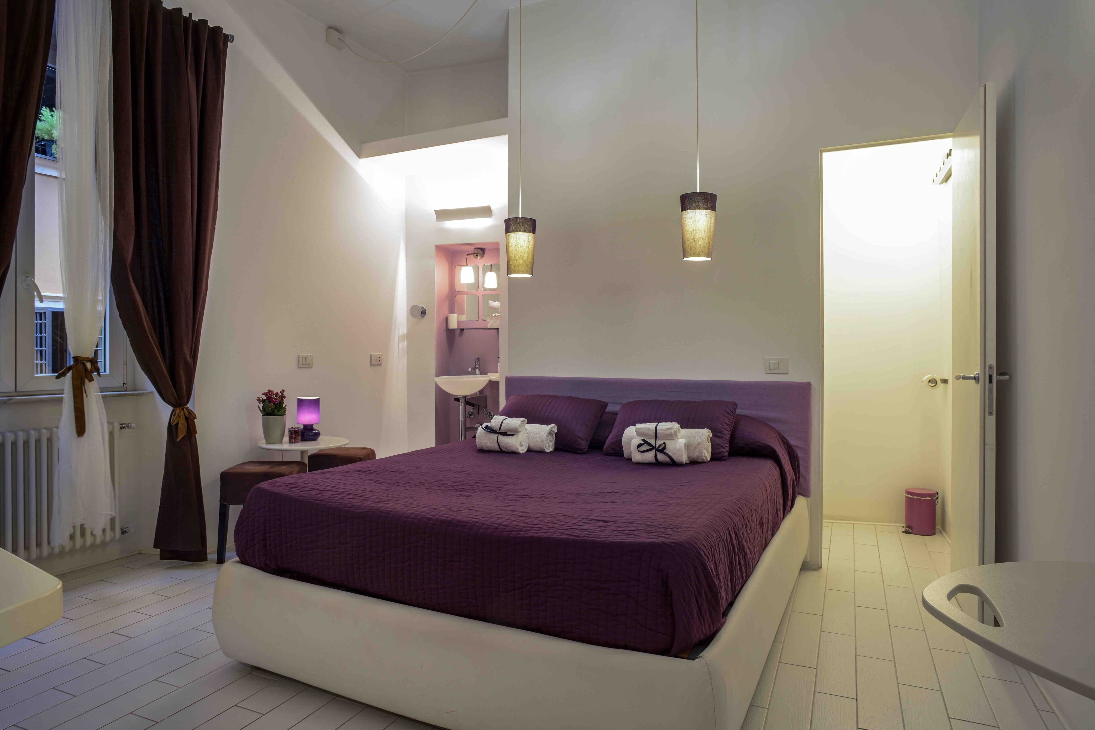 Une chambre à coucher moderne avec un grand lit, une table de chevet, une lampe et une salle de bain visible à travers une porte ouverte.