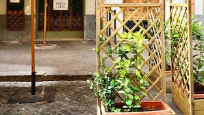 A street-side pizza and grill establishment with a wooden pergola, potted plants, and a traditional wooden door.