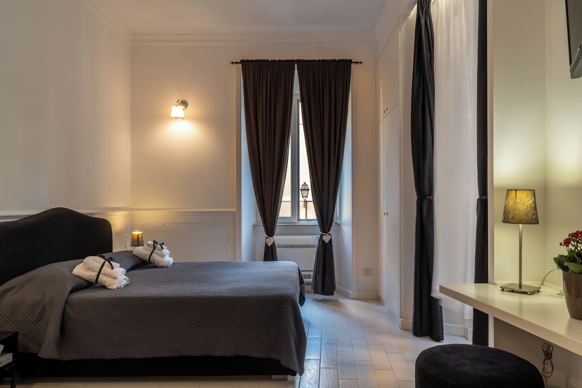 Une chambre d’hôtel avec un lit, une table de chevet avec une lampe, une fenêtre avec des rideaux et un luminaire fixé au mur.