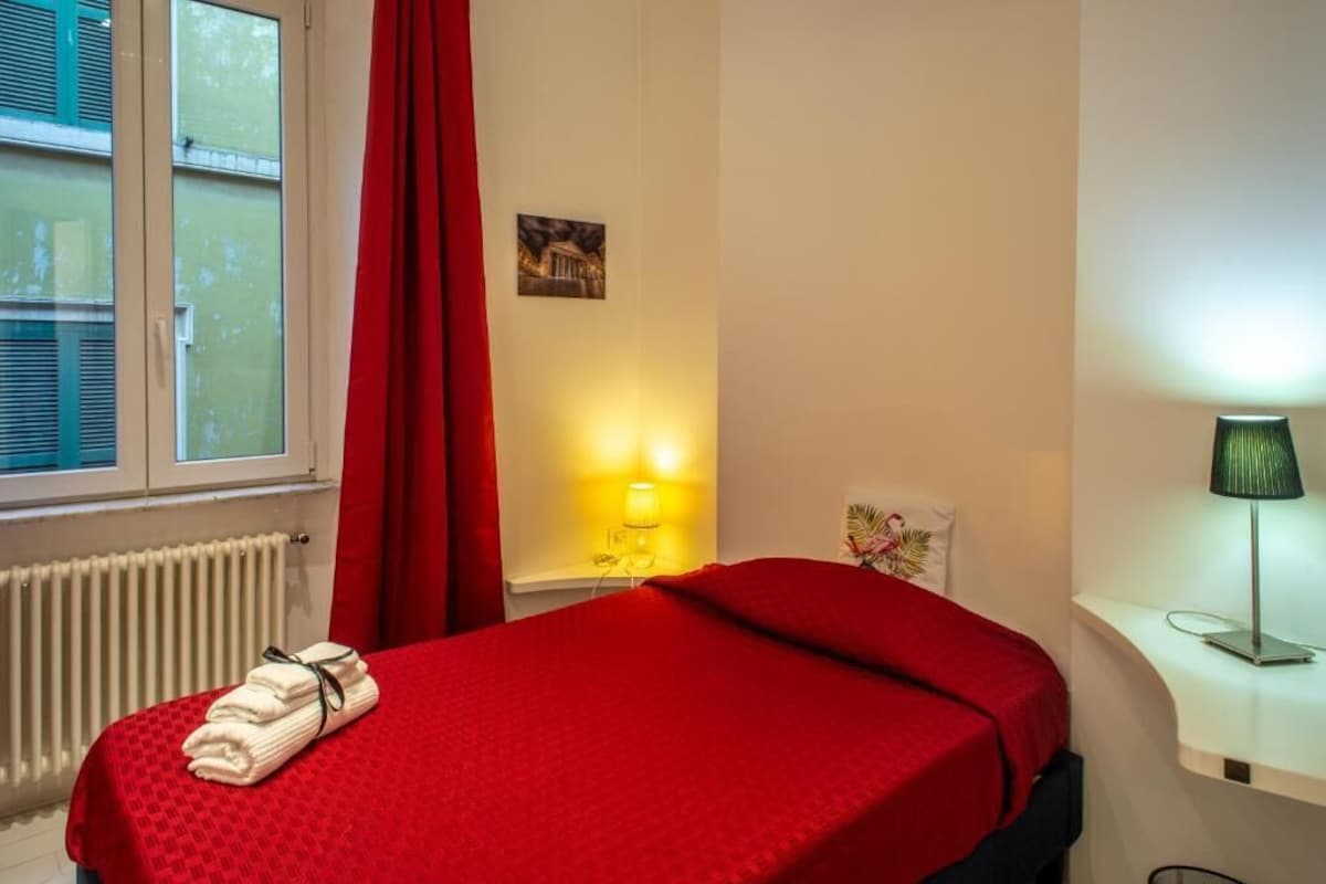 Une chambre d’hôtel avec un lit, un radiateur, une fenêtre avec des stores, un rideau rouge, une lampe fixée au mur et un petit tableau.