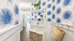Un couloir avec un mur blanc orné de motifs de feuilles de palmier bleues, une console blanche surmontée d’un panier en osier et une étagère blanche agrémentée d’objets décoratifs.