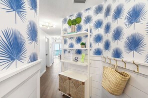 A hallway with a white wall featuring blue palm leaf patterns, a white console table with a woven basket, and a white shelving unit with decorative items. - 17th Floor Gulf-Front Luxury | 2 Baths | Beach Service Included (Panama City Beach)