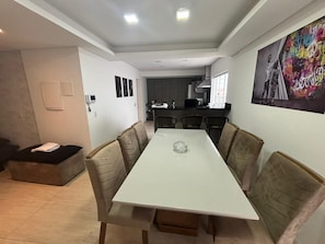 A modern dining area with a white table and cushioned chairs, a kitchen in the background, and a colorful artwork on the wall.