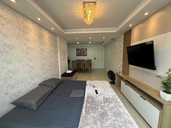 A modern hotel room with a bed, a TV, a desk, and a dining area in the background.