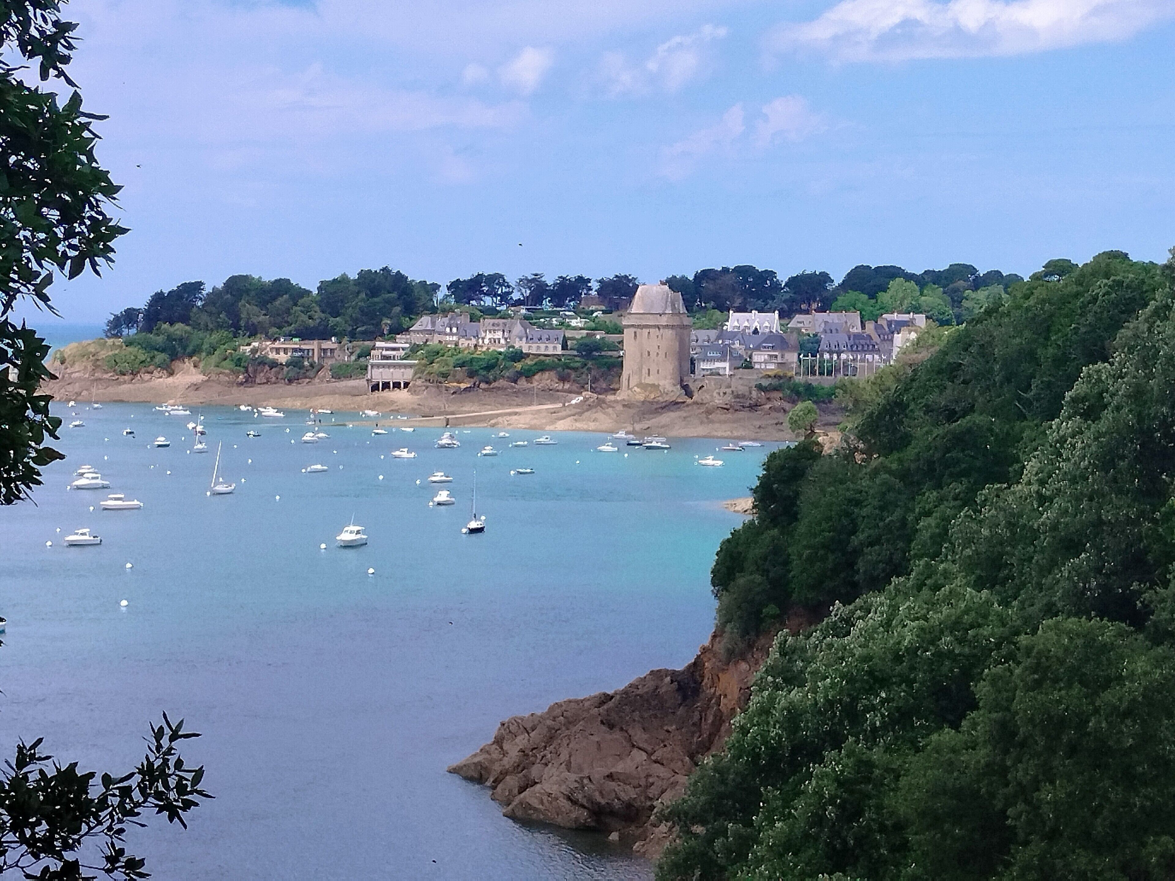 A coastal town with a prominent tower, boats on the water, and lush greenery.