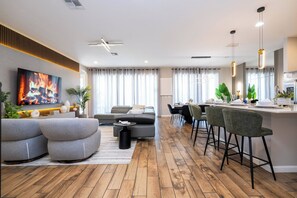 A modern living room with a sofa, a coffee table, and a TV mounted on the wall. There is a dining area with a table and chairs, and a kitchen area with a bar counter and stools. - DLuxx Luxury Themed Home / Pool / Near Disney 438 (Kissimmee)