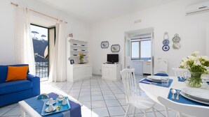 Un salón luminoso con un sofá azul, una mesa de comedor para dos personas y un televisor sobre un mueble blanco.