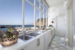 Cozinha com vista para o mar, armários brancos, pia e cesta de legumes.