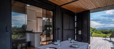 An open-plan kitchen with a dining area on a wooden deck, overlooking a mountainous landscape.