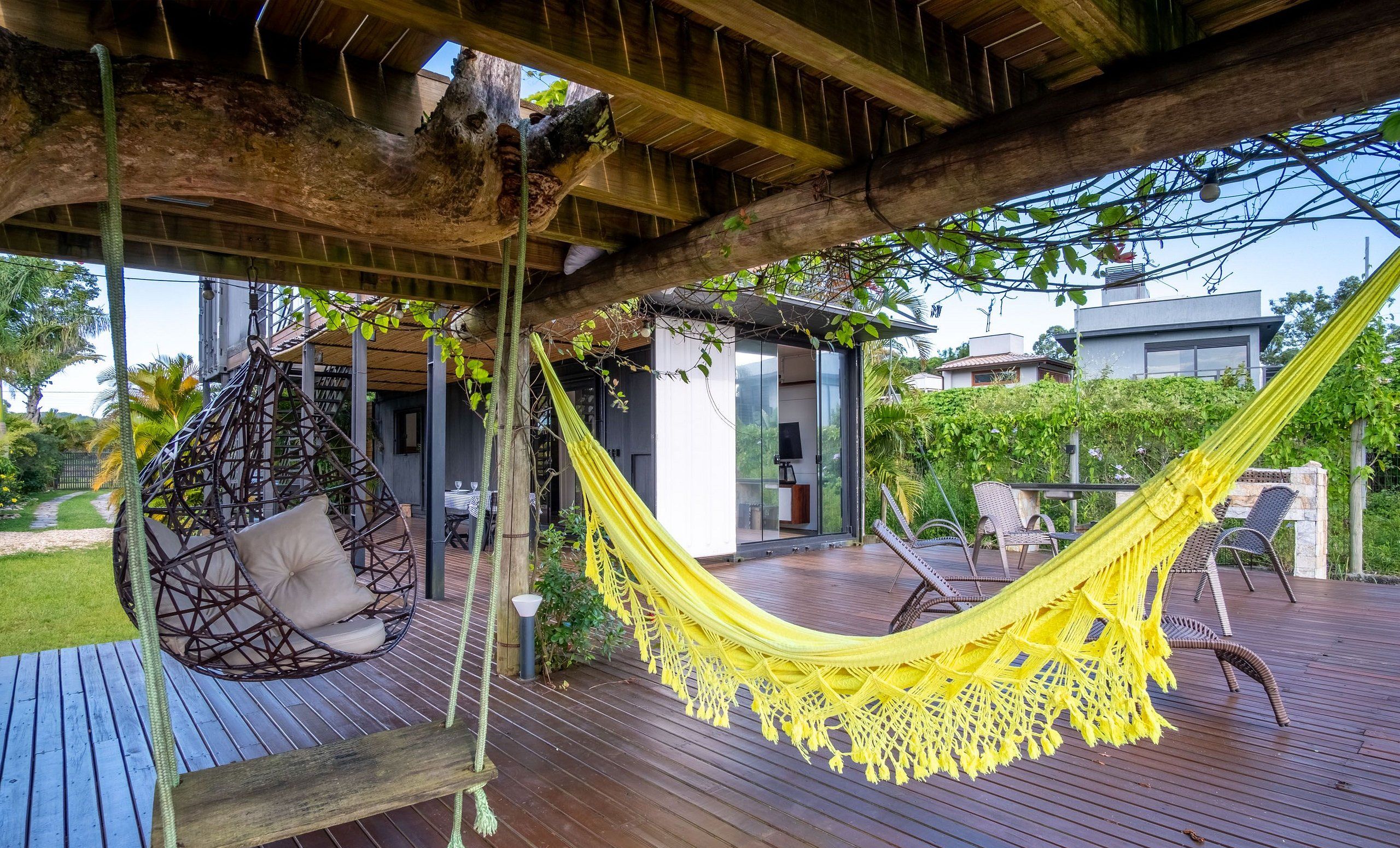 A hammock and a hanging chair on a wooden deck with a view of a modern house and greenery.