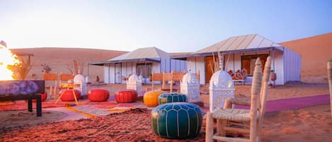 Un campamento en el desierto con tiendas de campaña, asientos coloridos y una fogata.