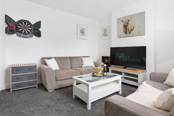 Ein modernes Wohnzimmer mit einem grauen Sofa, einem Couchtisch, einem Fernseher und einer Dartscheibe.