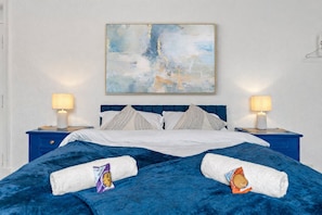 Ein Bett mit blauer Decke, zwei Kissen und zwei Handtüchern. Darüber hängt ein Gemälde, und an beiden Seiten des Bettes stehen Nachttischlampen.