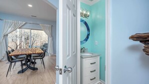 Un comedor con una mesa y sillas de madera, vista a árboles a través de un ventanal grande, y un baño con lavamanos y un espejo decorativo.