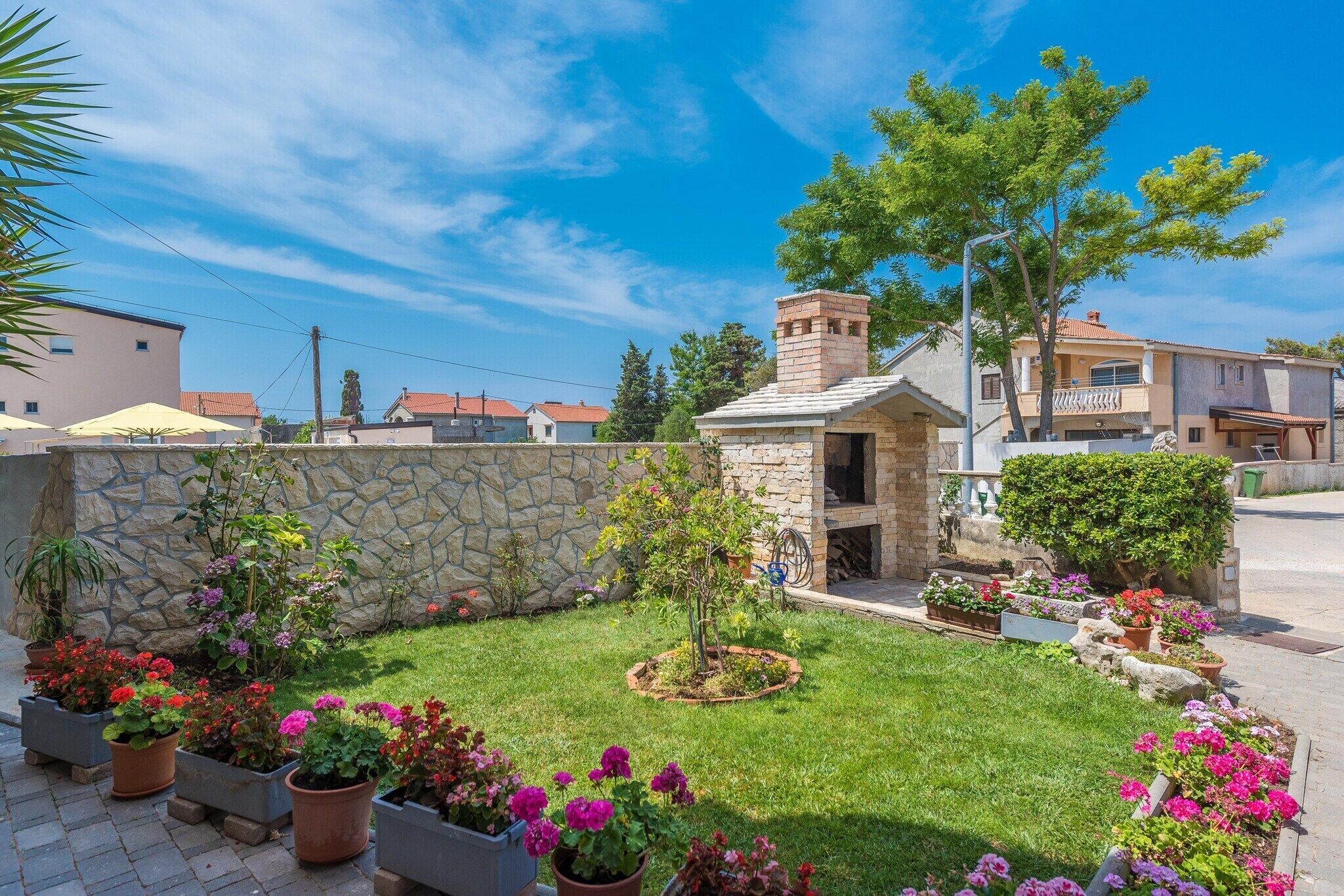 Un giardino curato con diverse piante fiorite e un piccolo albero, circondato da mura in pietra e un camino in pietra.