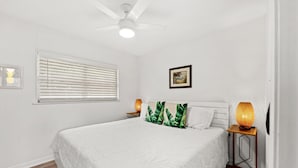 Un dormitorio con cama, ventilador de techo, ventana con persianas, lámpara de noche y un cuadro en la pared.