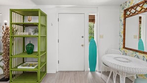 Una habitación con una puerta blanca, un estante verde con objetos decorativos, una mesa de mimbre blanca y un espejo con marco de bambú.