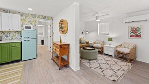 Cocina moderna con gabinetes verdes, un refrigerador azul claro y un carrito de madera. Hay un comedor con mobiliario de mimbre y una alfombra estampada.