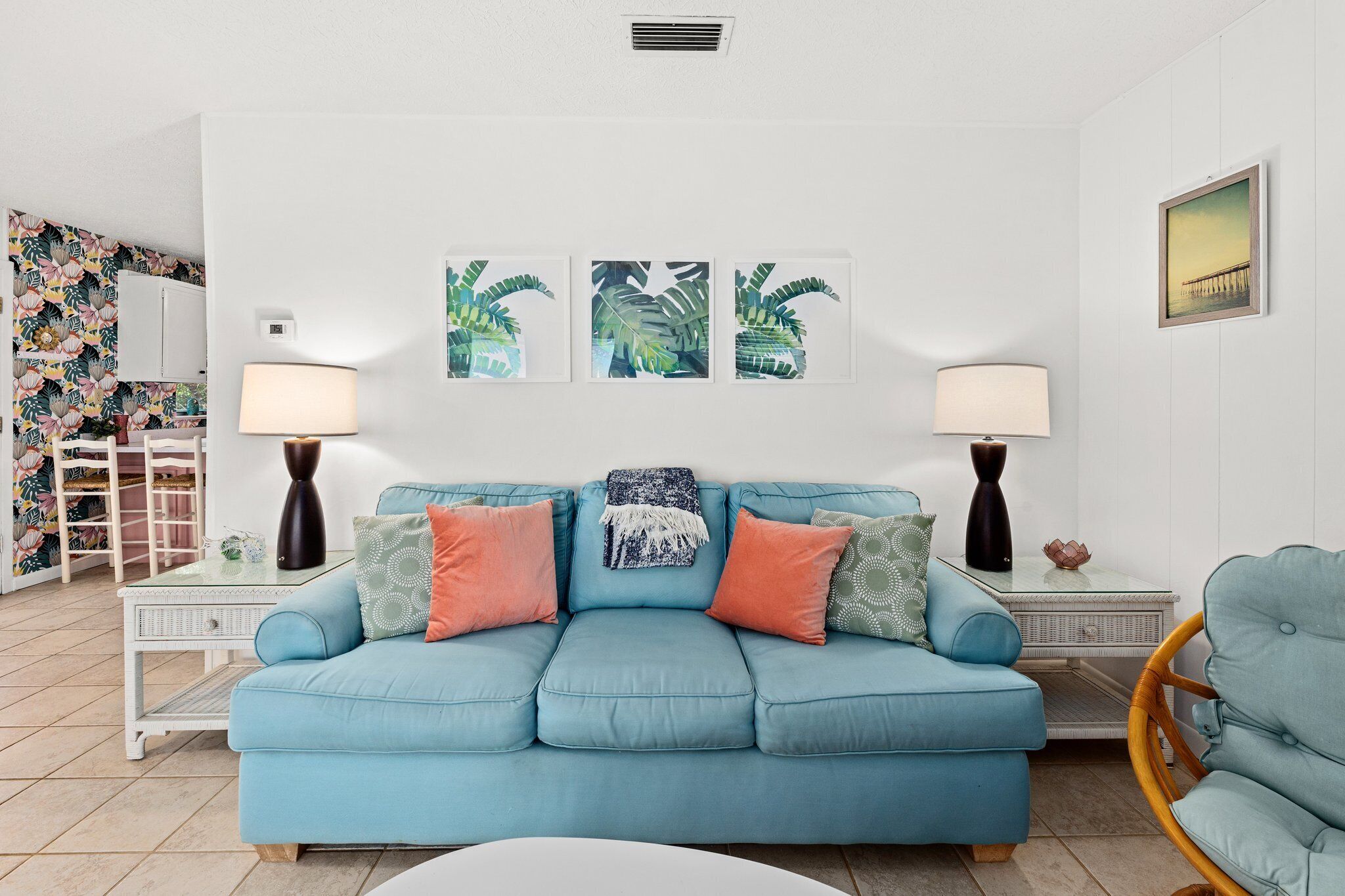 Un salón con un sofá azul, dos mesitas de noche con lámparas, un sillón de mimbre y una pared con tres cuadros enmarcados.