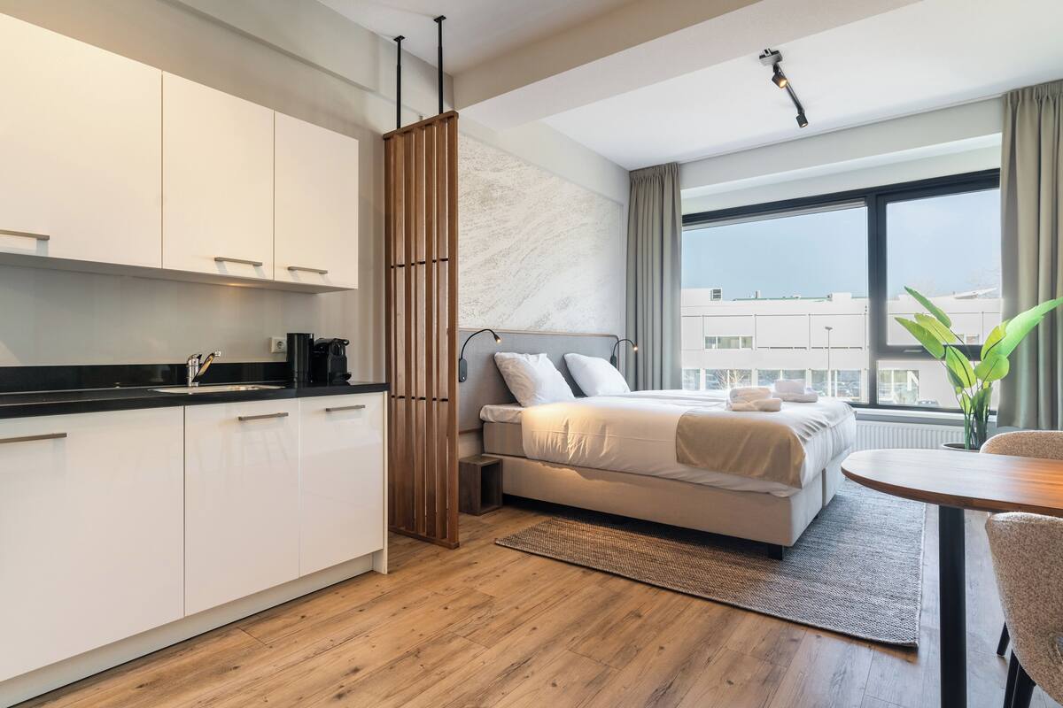 A modern hotel room with a kitchenette, a bed with a view, and a dining area.