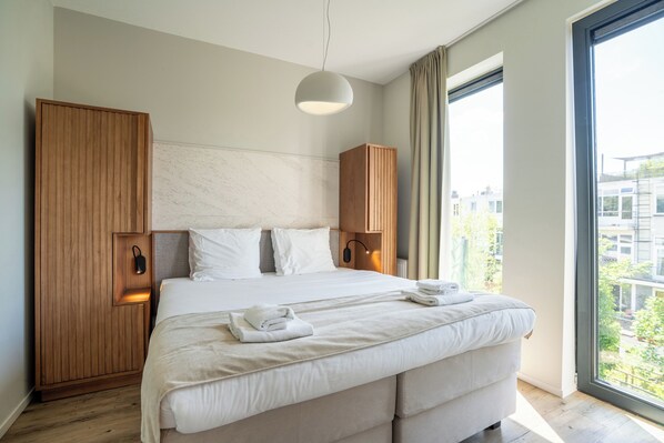 A modern hotel room with a large bed, wooden wardrobe, and a view of the city through large windows.
