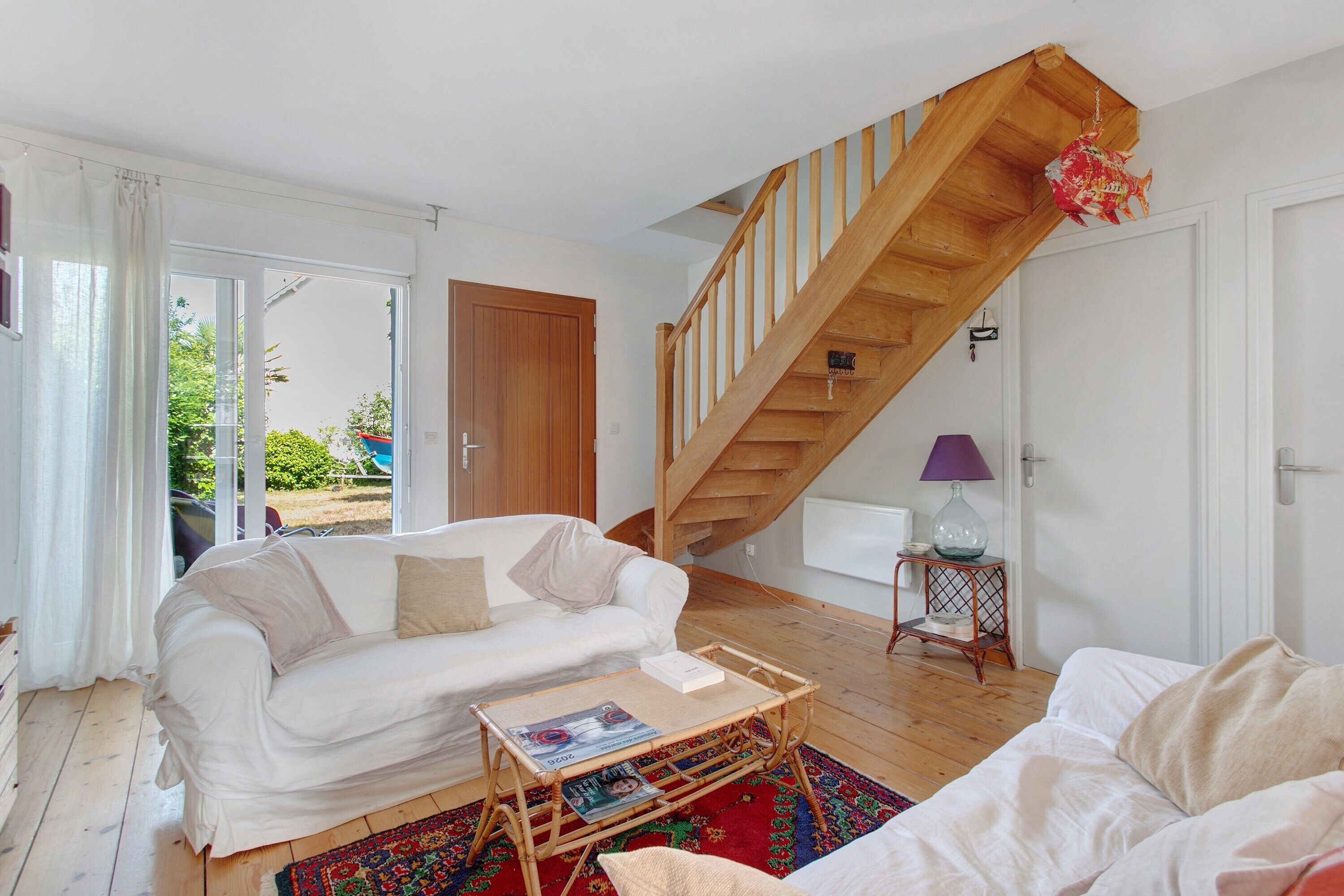 Een woonkamer met een houten trap, witte meubels en uitzicht door een glazen deur.