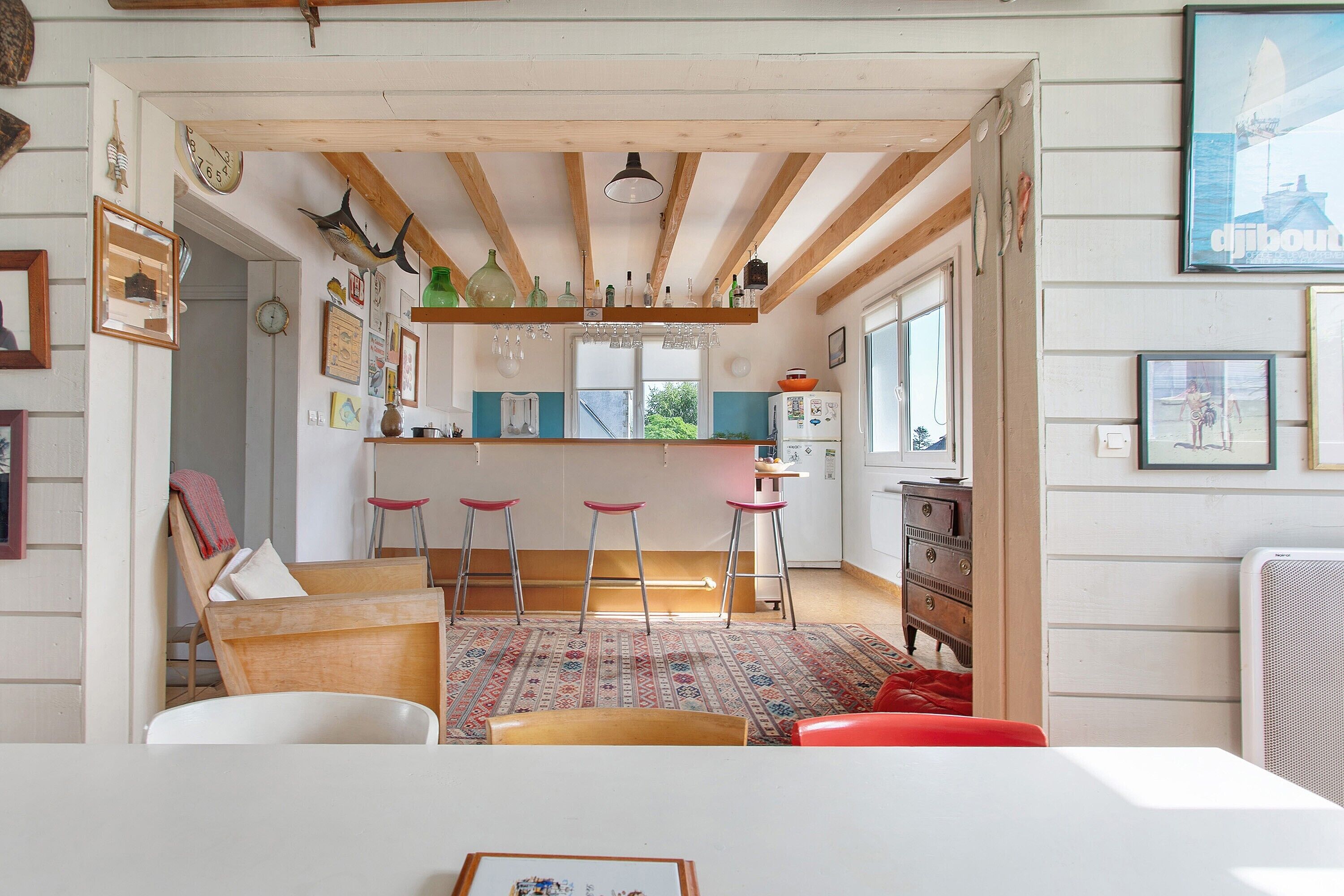 A room with a wooden ceiling, a kitchen area with bar stools, a patterned rug, and various framed pictures on the walls.