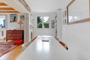 Ein langer Esstisch mit Stühlen, eine hölzerne Kommode, ein roter Sitzsack und ein Fenster mit Blick auf Grünflächen.