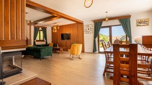A spacious living area with a wooden dining table, a green sofa, and a yellow armchair. A flat-screen TV is mounted on the wall, and there is a fireplace on the left.