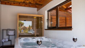 A room with a hot tub, two glasses of wine, towels on a shelf, and a window with wooden shutters.