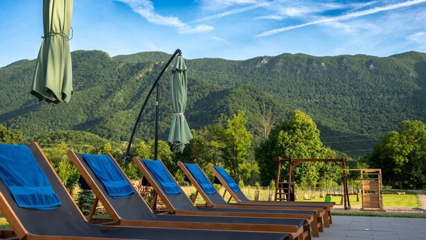 A row of wooden lounge chairs with blue towels, a wooden swing set, and a view of green mountains under a clear sky.