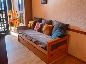 Un sofá de madera con cojines, un banco de madera y una mesita en una habitación con suelo de madera y una pared blanca con textura.