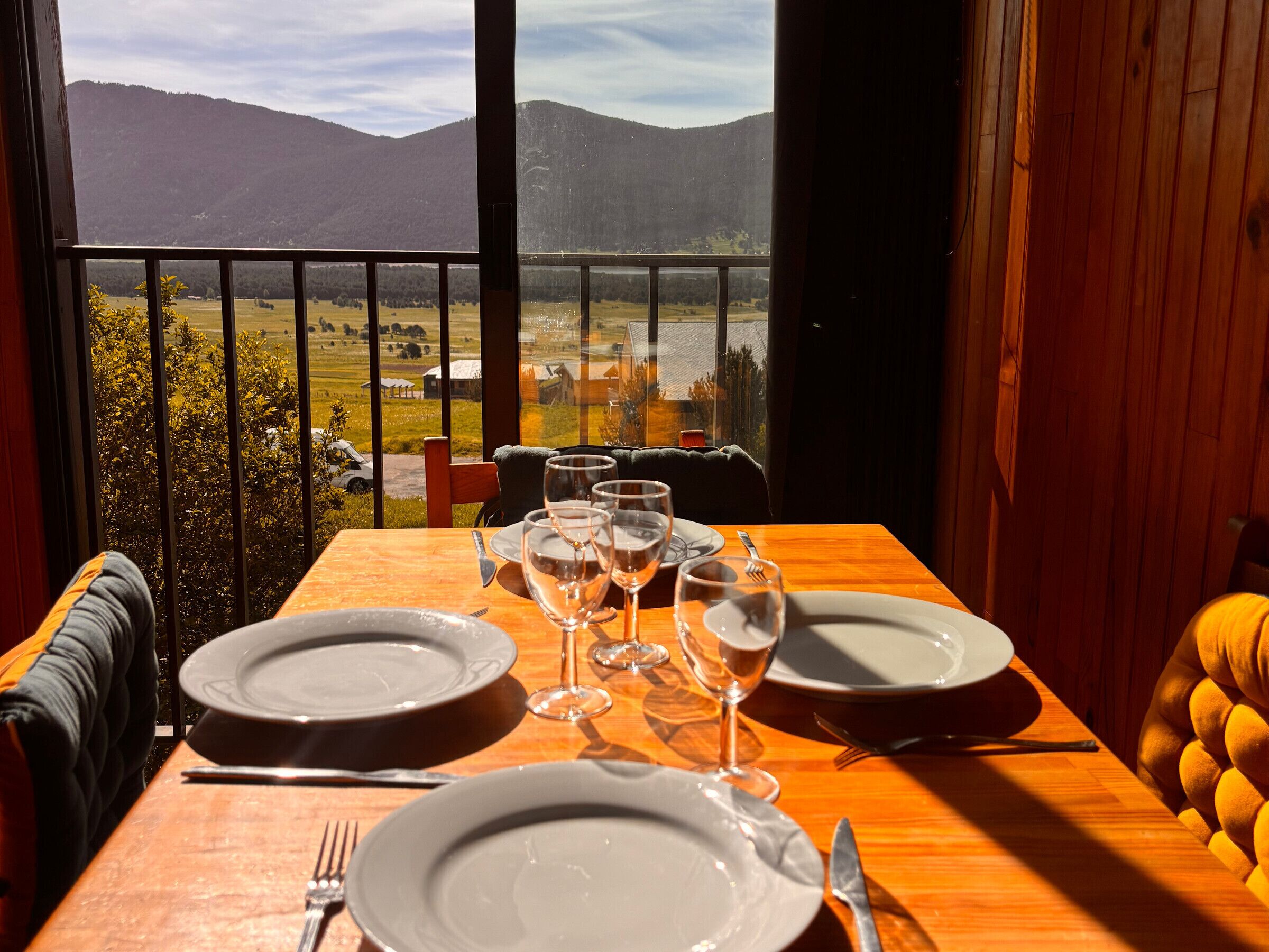 Mesa de jantar posta com pratos, copos de vinho e talheres, com vista para uma paisagem montanhosa.