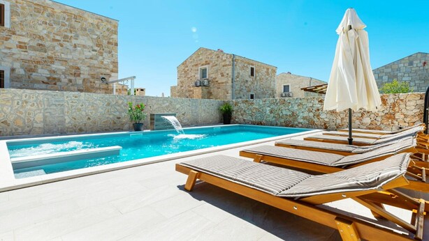 Una piscina con lettini e un ombrellone, sullo sfondo edifici in pietra e una cascata.