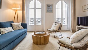 Uma sala de estar com um sofá azul, uma poltrona de vime, uma mesa de centro de madeira e uma televisão sobre um móvel.