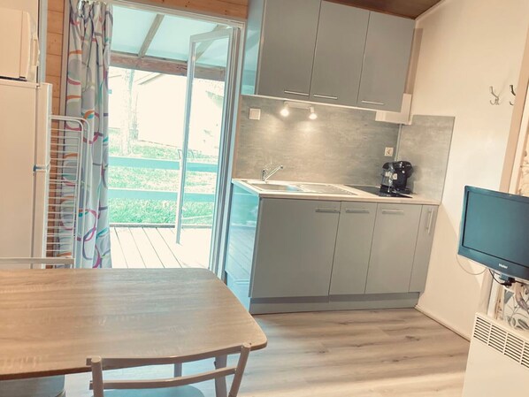A compact kitchen with grey cabinets, a sink, and a coffee maker. There's a wooden table and a TV mounted on the wall.