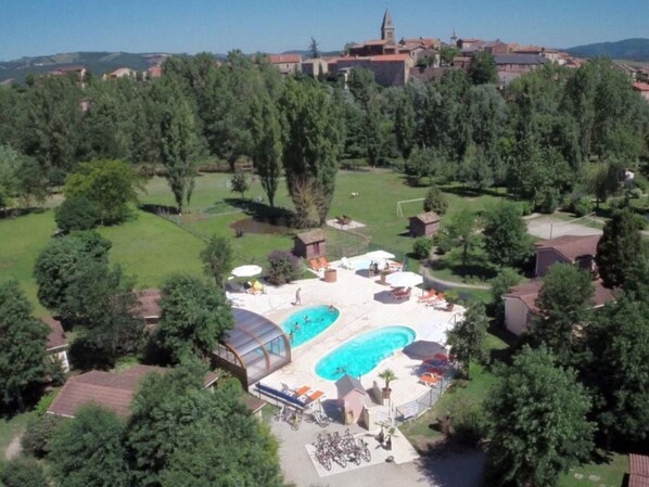 Vista aérea de un resort con dos piscinas, rodeado de exuberante vegetación y un edificio grande al fondo.