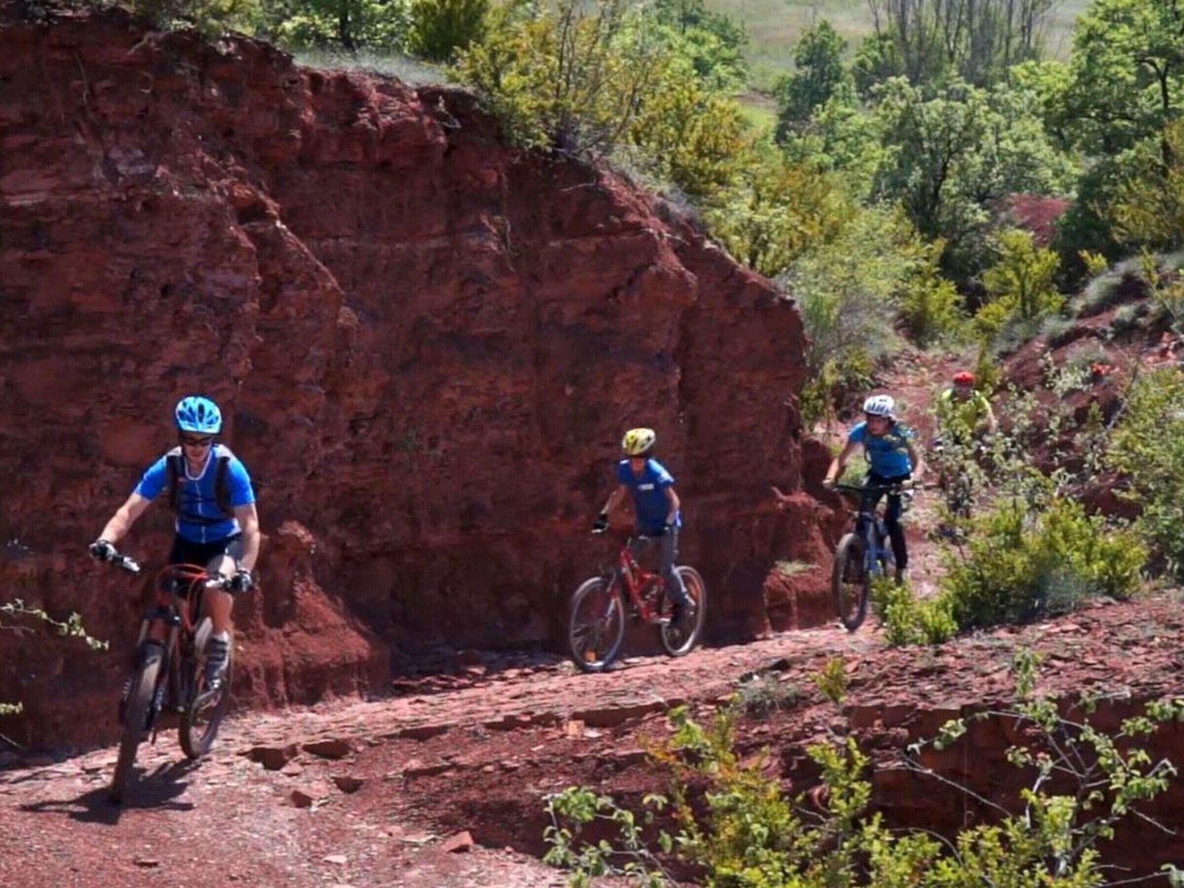 Three mountain bikers on a dirt trail with red rock formations and green vegetation in the background.