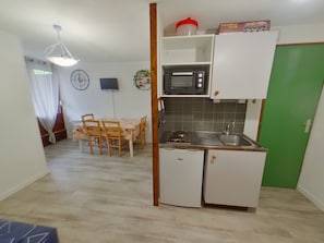 Eine kompakte Küche mit Mikrowelle, Spüle und kleinem Kühlschrank. Ein Esstisch mit Stühlen und eine Uhr sind an der Wand angebracht.