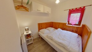 A small, neatly arranged bedroom with a single bed, a bedside table with a lamp, a shelf with toys, and a window with a red curtain. - Apartment in Les Rousses with Terrace (Les Rousses)