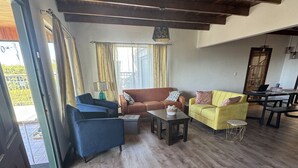 Amplia sala de estar con piso de madera, un sofá marrón, un sofá amarillo y un sillón azul. En el fondo, se observa una mesa de comedor con sillas.