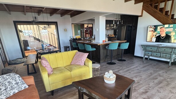 Un salón amplio con un sofá amarillo, una mesa de centro de madera y un otoman gris. Se ve una cocina con barra y taburetes al fondo.