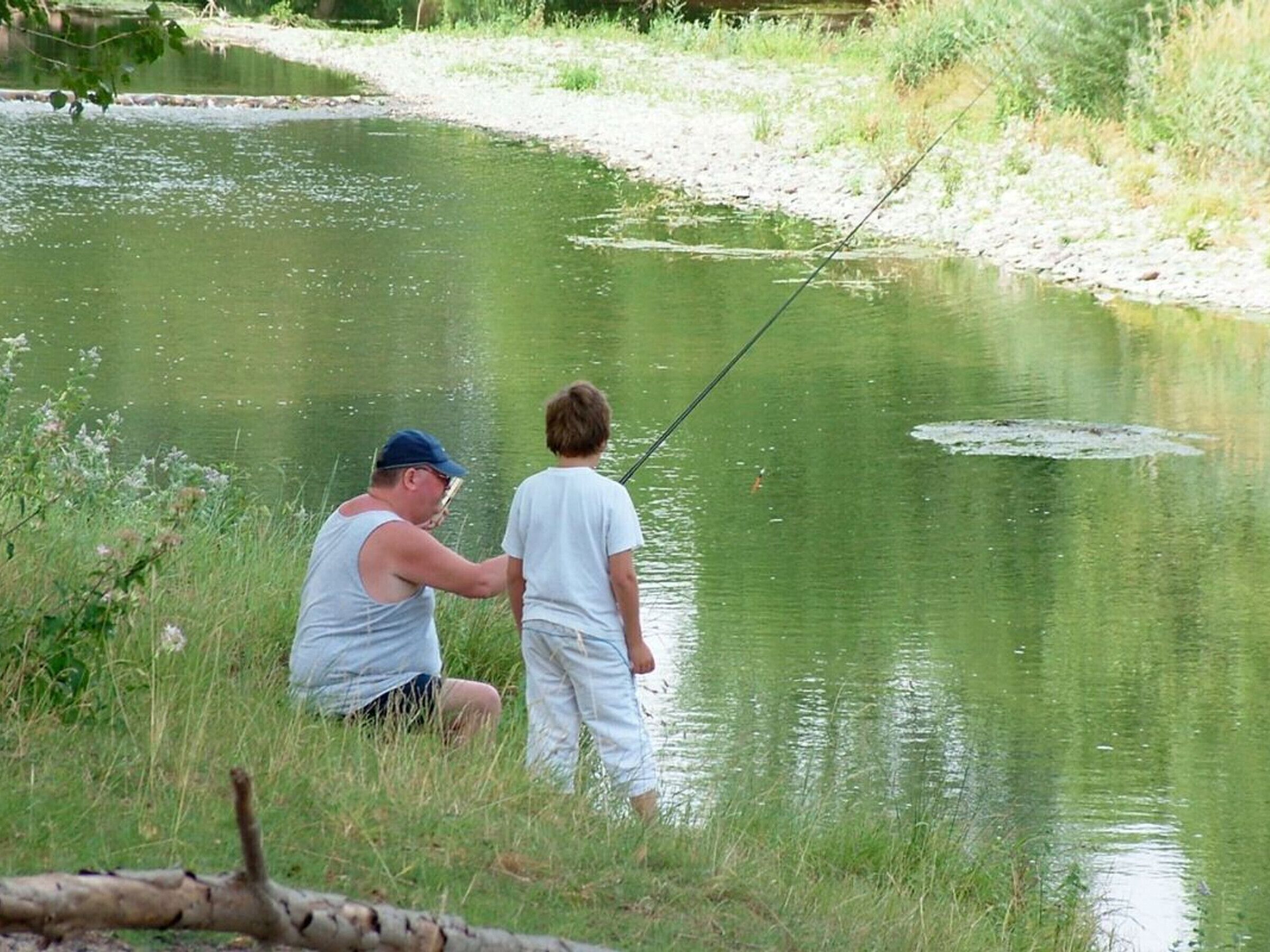 Two people fishing by a calm river surrounded by greenery.
