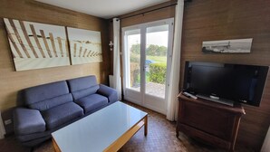 Een woonkamer met een bank, een salontafel, een televisie op een houten standaard en een glazen schuifdeur die naar een buitenruimte leidt.