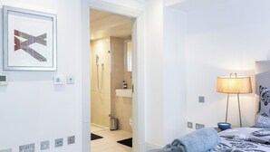 Una habitación de hotel moderna con una cama, una lámpara de noche, una obra de arte enmarcada y un baño visible a través de una puerta abierta.