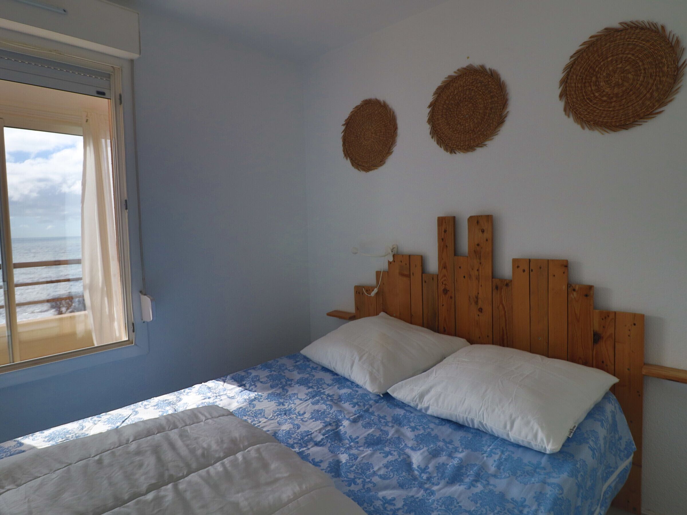 A bedroom with a wooden headboard, a bed with blue floral bedding, two pillows, and a window with a view of the sea.